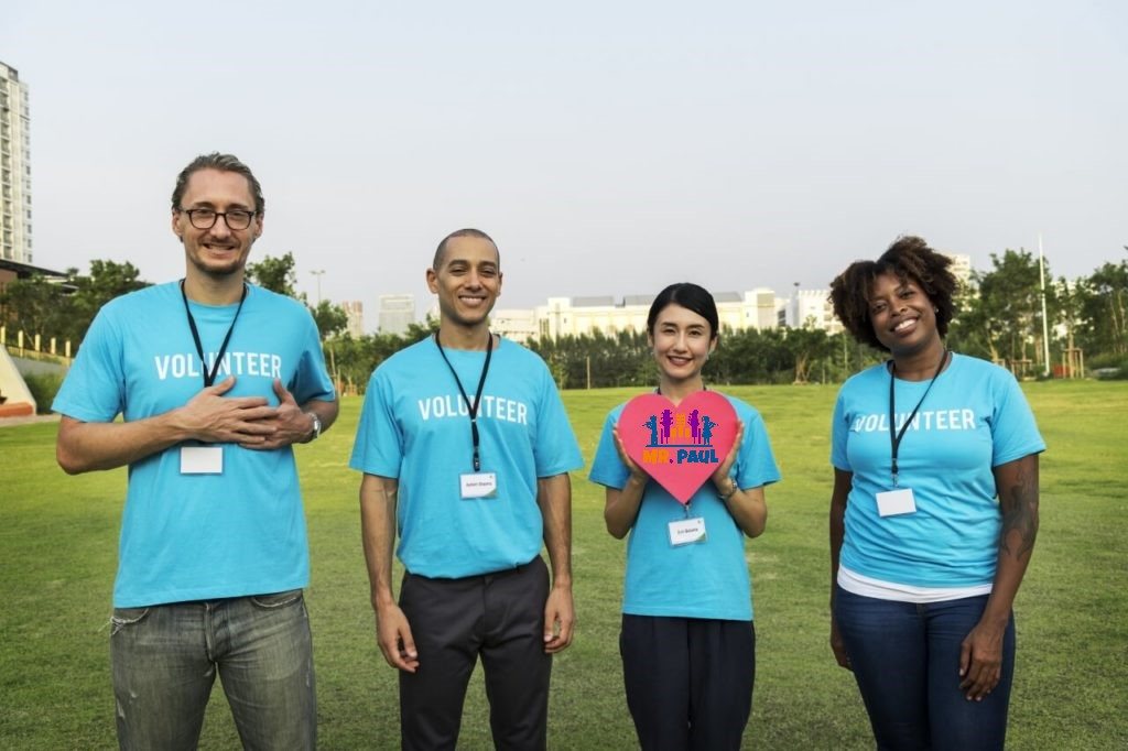Four Mr. Paul Foundation volunteers in matching blue shirts holding a heart-shaped Mr. Paul sign